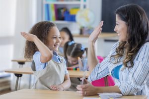 Happy school teacher gives young schoolgirl a high five after getting a math question correct. The teacher is using flashcards to help the young student.